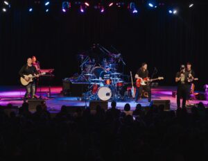 A band performs on stage with guitars, drums, and keyboards under colorful stage lights.