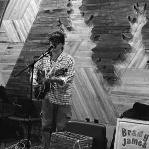 Shane Hardwicke, in glasses and cap, plays guitar and sings into a mic on a wooden stage.