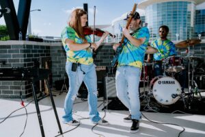 Three musicians in tie-dye shirts performing outdoors with violin, guitar, and drums.