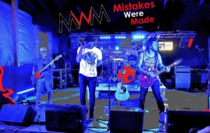 A rock band performs onstage under blue and purple lights with Mistakes Were Made behind them.