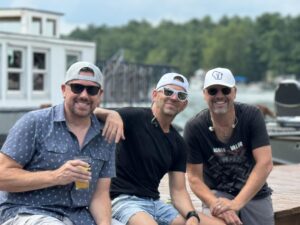 Three men in sunglasses and hats sit smiling outdoors by water, one holding a drink.