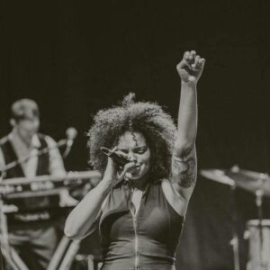 A singer with curly hair performs passionately on stage with one arm raised and eyes closed.
