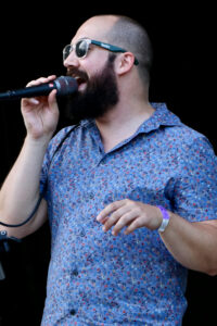 A bearded man in sunglasses sings into a microphone while wearing a blue floral shirt.