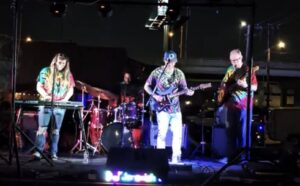 Four musicians in tie-dye shirts perform on an outdoor stage at night with instruments and lights.