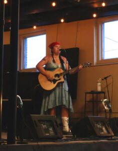 Person in a red bandana plays acoustic guitar and sings on an indoor stage with string lights.