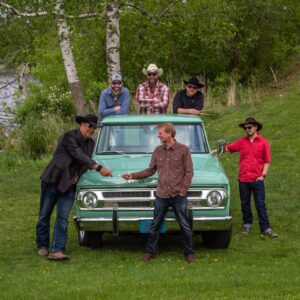 Six men in casual western wear pose around a vintage green truck on a grassy field.