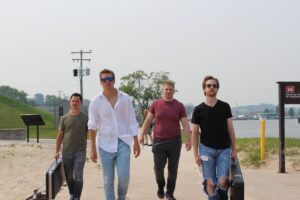Four men walk outdoors carrying instrument cases on a sunny day by the water.