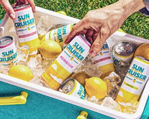 Hands grabbing Sun Cruiser lemonade vodka cans from an ice-filled cooler with lemons inside.
