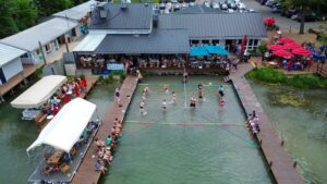 Aerial view of people playing volleyball in the water next to a lakeside restaurant with docks.