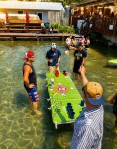 Four men play beer pong on a floating table in shallow water, with others relaxing nearby.