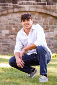 Young man in white shirt and jeans kneels on grass, smiling, with stone wall in background.