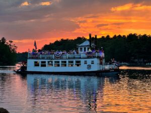 A white riverboat with passengers sails on a lake at sunset, decorated with patriotic bunting.